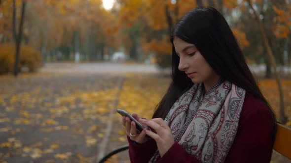 Attractive Woman Relaxing in Autumn Park Sitting on Bench and Using Smartphone