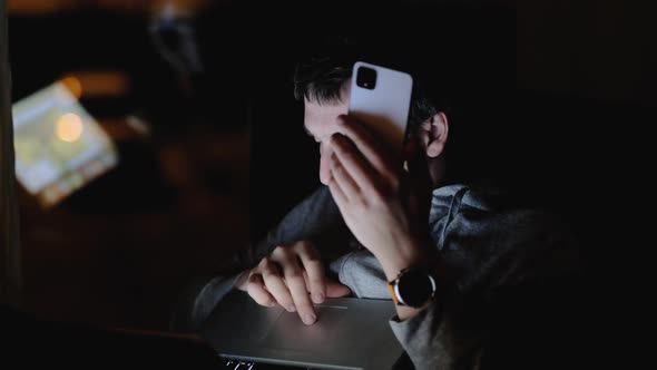 Man Surfs Internet with Cellphone and Laptop in Dark Room