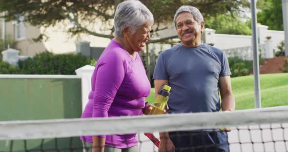 Video of happy biracial senior couple holding water and tennis balls on tennis court