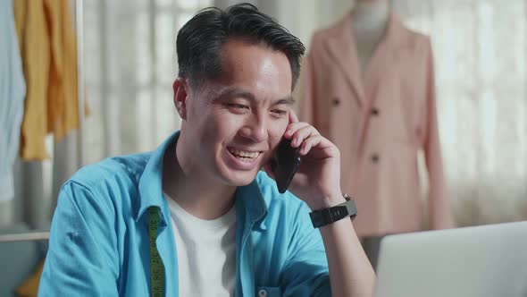 Close Up Of Asian Male Designer With Sewing Machine Talking On Smartphone While Designing Clothes 