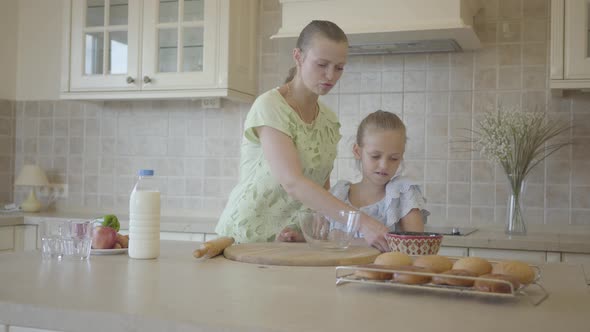 Mother Helping and Teaching Her Little Cute Daughter in Modern Cozy Kitchen To Cook Cake or Pancakes