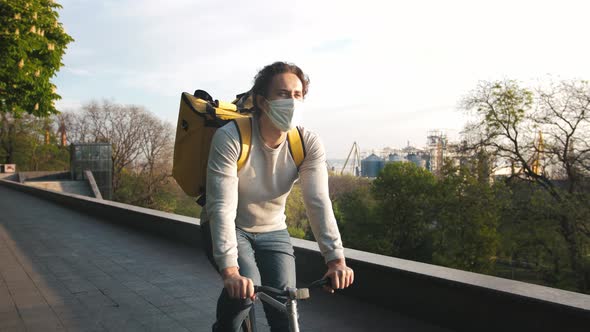 Portrait of a Courier with Yellow Backpack and Protective Mask Rides a Bicycle Through the City Park