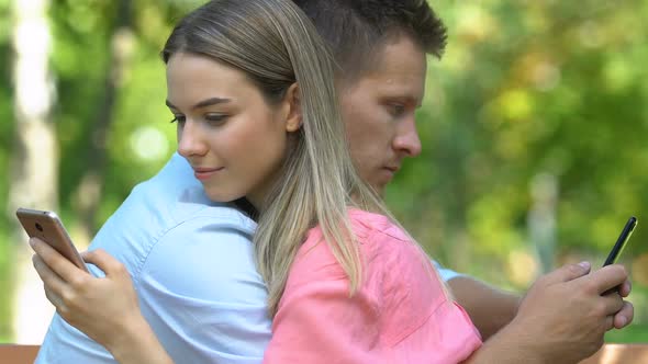 Couple Tenderly Hugging Sitting on Bench and Chatting on Smartphones, Close-Up
