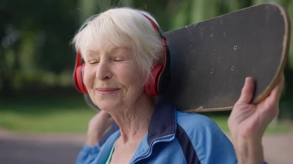 Closeup Cheerful Beautiful Active Senior Woman in Headphones with Skateboard on Shoulders Smiling