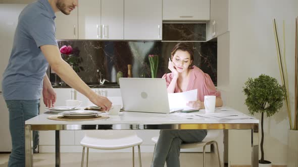 An Adult Woman is Sitting and Working at Home While Her Husband is Doing Household Chores