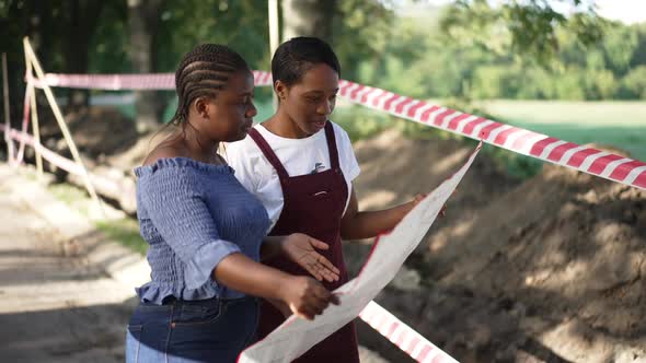 Two Confident Professional African American Women Standing with Blueprint in Summer Park at