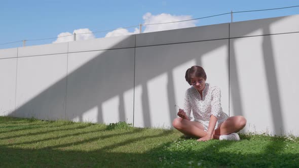 Woman with Colorful Sunglasses Sits on Lawn in Urban Park and Enjoys of Bright Sunlight