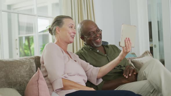 Happy senior diverse couple wearing shirts and using tablet in living room