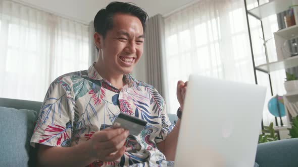 Happy Asian Man Sitting On Couch and Holding Credit Card And Using Laptop Computer Shopping Online