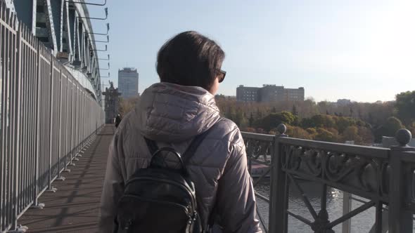 A Middleaged Woman Walks Alone with a Bouquet of Yellow Leaves on a Bridge Over a River on a Sunny