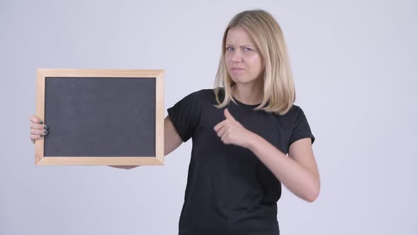 Young Serious Blonde Woman Holding Blackboard and Giving Thumbs Down