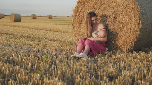 Beautiful Young Mother Feeds Baby Son with Breast at Haystack in Summer Field