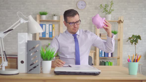 Man in a Shirt Sitting at a Laptop in the Office with a Piggy Bank