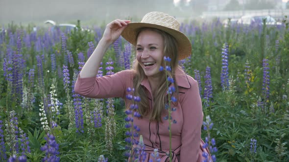 Girl in Pink Dress Rises From Grass in Blooming Lupine Field and Waving Boater with Straw Hat at