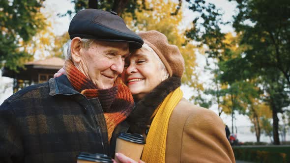 Elderly Husband and Wife Holding Paper Cups of Coffee Smiling and Hugging Posing in Autumn Park