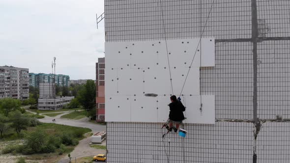 Aerial View on Industrial Climber Suspended on Ropes Performs Work on Insulation of Facade of High