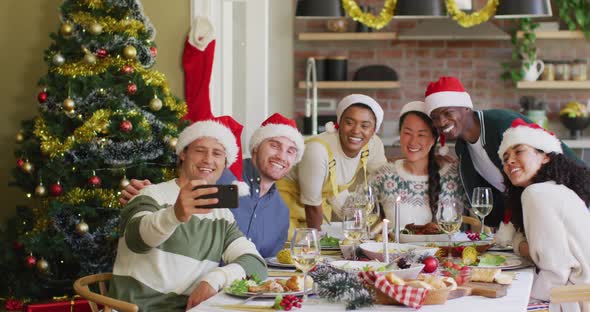 Happy group of diverse friends in santa hats celebrating meal, taking selfie at christmas time