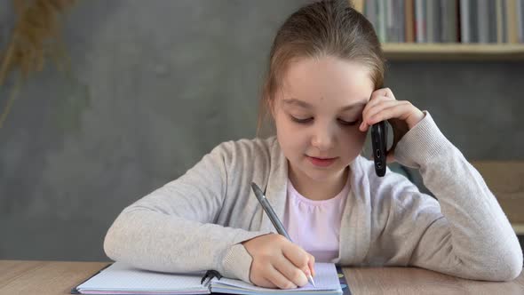 Schoolgirl Girl Talking on the Phone and Making Notes in a Notebook Sitting at Home at the Table