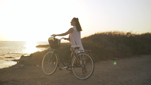 Side View Portrait of Slim Young Woman Strolling with Bike in Sunbeam at Sunset on Cyprus
