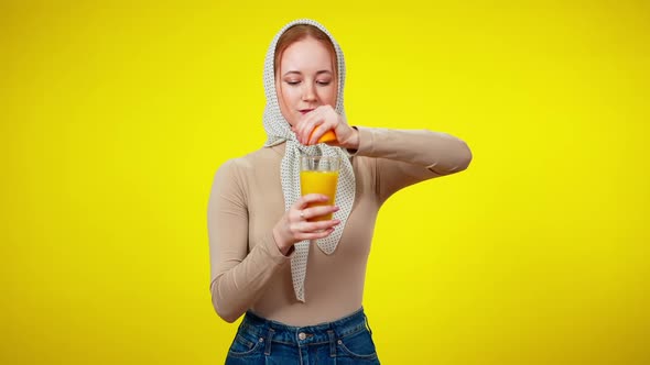 Smiling Happy Redhead Woman in Kerchief Squeezing Orange in Juice Glass at Yellow Background