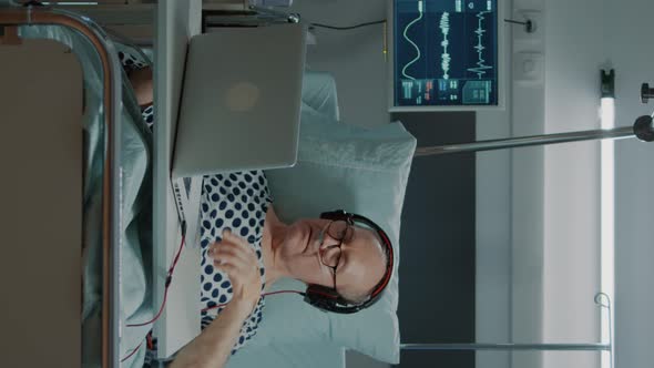 Vertical Video Sick Old Man Sitting in Hospital Ward Using Laptop and Headphones