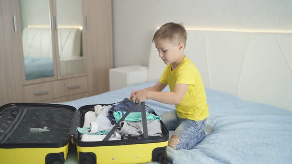 Happy Child Closes a Suitcase While Sitting on the Bed in the Bedroom