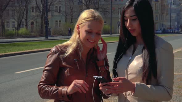 A Young Caucasian Woman and a Young Asian Woman Listen To Music on a Smartphone in a Street