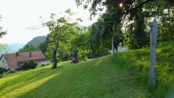 A man in shorts and green shirt is mowing a green meadow with a small lawnmower. Aerial 4k view.