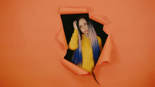 Thoughtful Young Woman Scratching Head and Looking Out of Hole of Orange Background