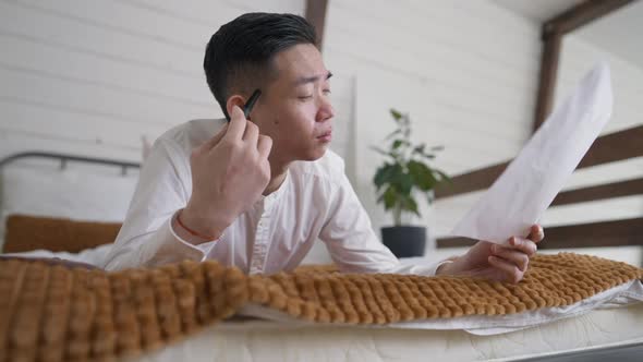 Focused Handsome Chinese Man Analyzing Documents Thinking Lying on Bed in Home Office