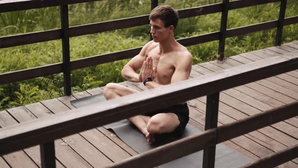 A calm half-naked guy with closed-eyes is doing meditation on the bridge