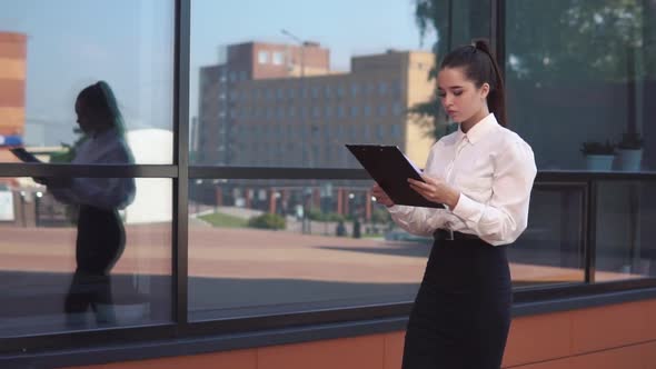 Young Business Girl with Dark Hair Goes Near Business Center Holding Documents in Hands