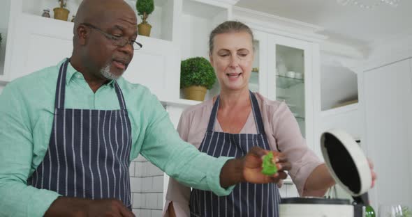 Smiling senior diverse couple wearing blue aprons and cooking in kitchen