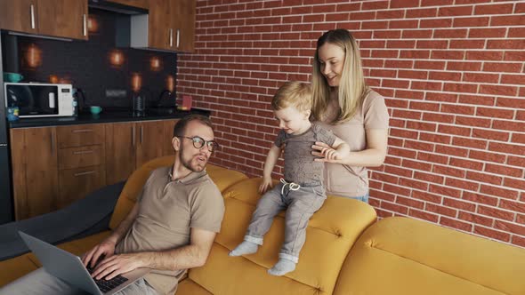 Mother Plays with Her Son Distracting Their Father Busy with Laptop on the Sofa