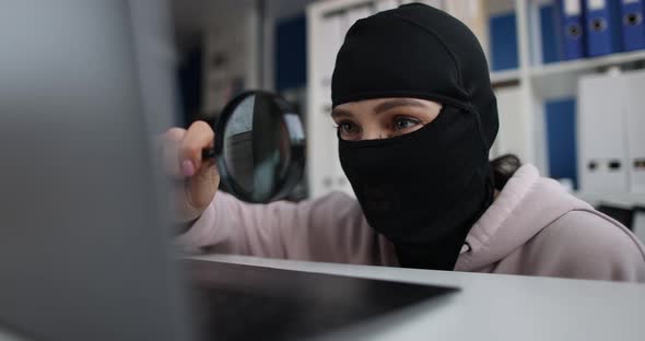 Swindler and Criminal in Balaclava Holds Magnifying Glass and Looks Computer