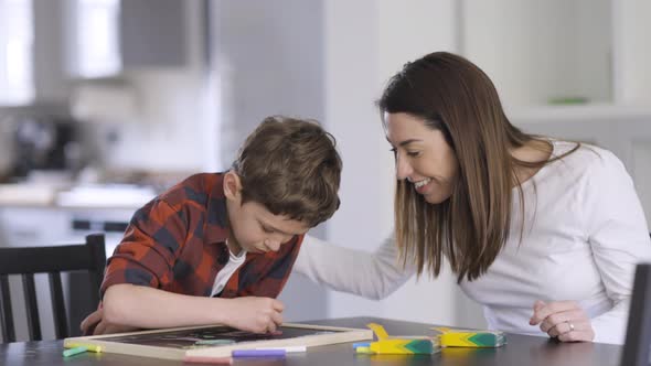 mother watching son painting with crayons on blackboard