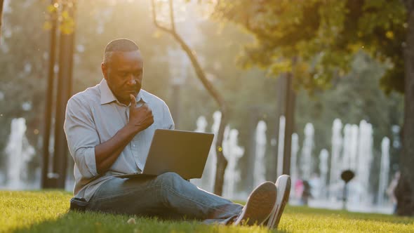 Serious Focused Senior Black Person Sitting on Grass in Park Using Laptop African American Mature