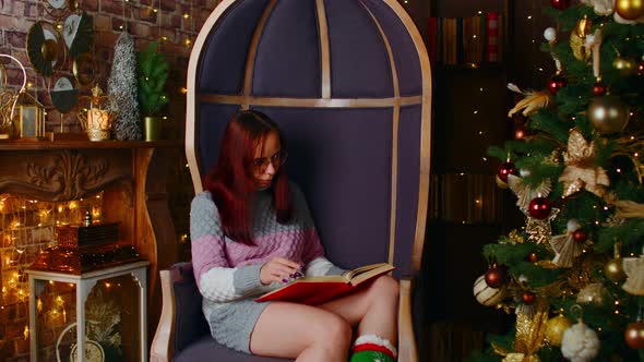 Concentrated Female in Warm Sweater and Eyeglasses Reading Book in Spare Time Near Christmas Tree