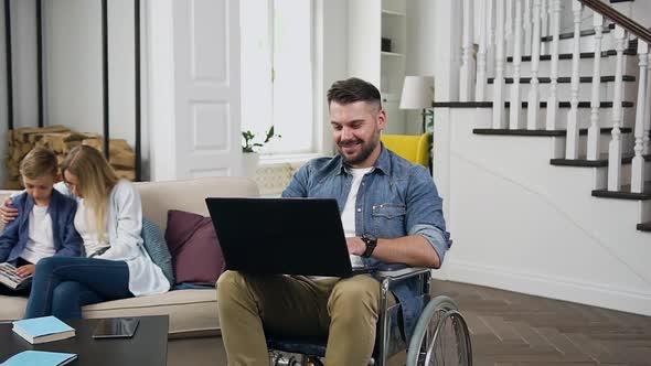 Bearded Guy in Wheelchair Working on Laptop and His Wife and Son Reading Book on the Sofa Near Him