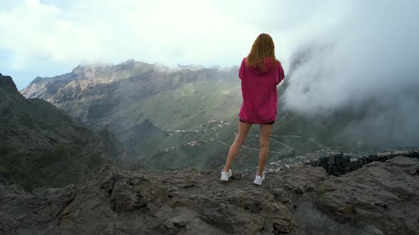 Beautiful Traveler Woman in a Pink Sweater Stands on the Edge of a Cliff and Enjoys the View of the