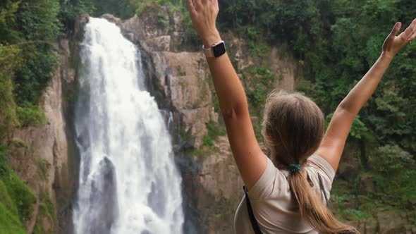 Young Happy Travel Woman Looking on Big Waterfall and Raised Her Hands Up