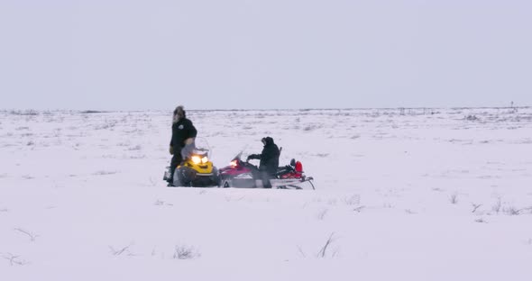 Wide shot of men on two snowmobiles in a snowy plain. The red snowmobile drives up to and past the s