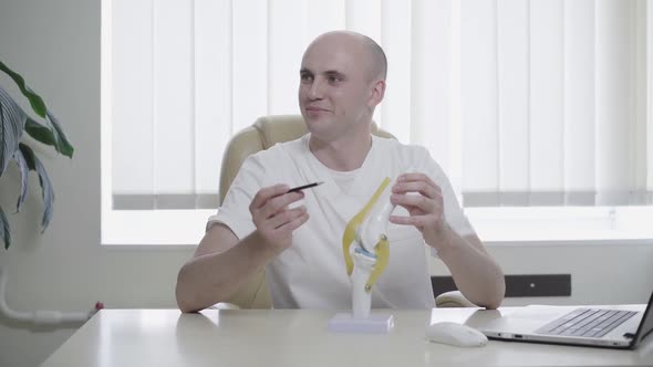 Front View of Confident Cheerful Traumatologist Sitting at the Table with Knee Joint Model and