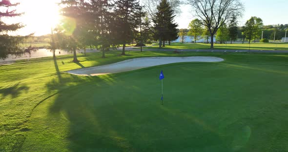 Rotating drone shot of golf course flag. Sun shines through trees onto beautiful green. Springtime i