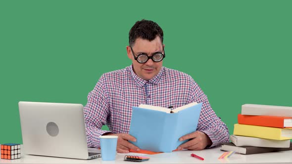 A Man in Glasses with with Thick Lenses Sits at a Desk in Front of a Laptop and Reads a Book Lifts