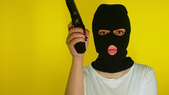 Unrecognizable woman in black balaclava with gun on yellow background.