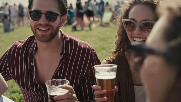 Group of friends sitting on grass together at music festival and drinking beer while chatting. Shot