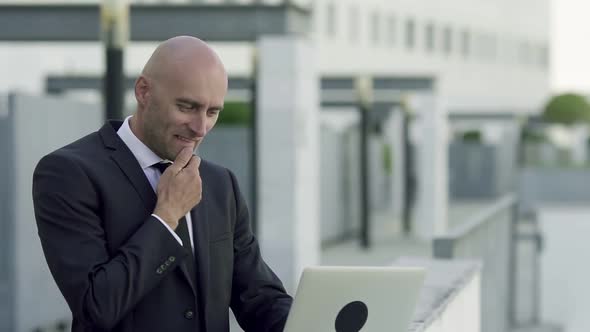 Concentrated Bald Office Employee Using Laptop and Rubbing Chin