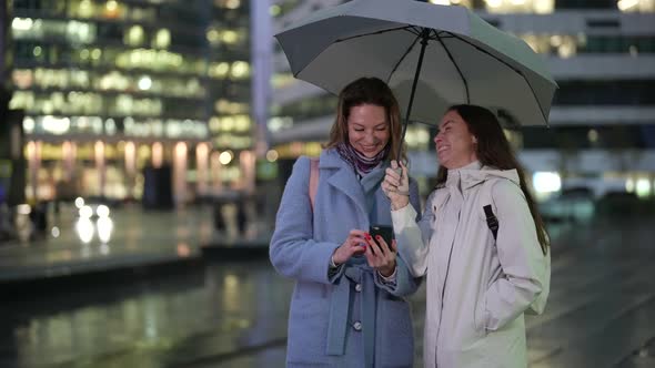 Ladies are Communicating on Street in Downtown in Rainy Evening Viewing Pictures in Mobile Phone