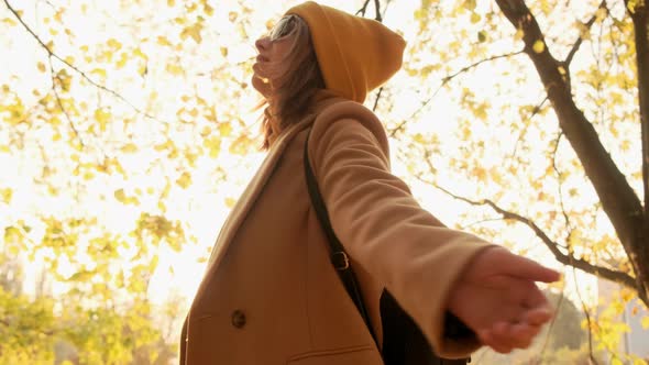 A Girl in a Brown Coat and an Orange Hat with a Backpack Enjoys the Suns Rays in an Autumn Park